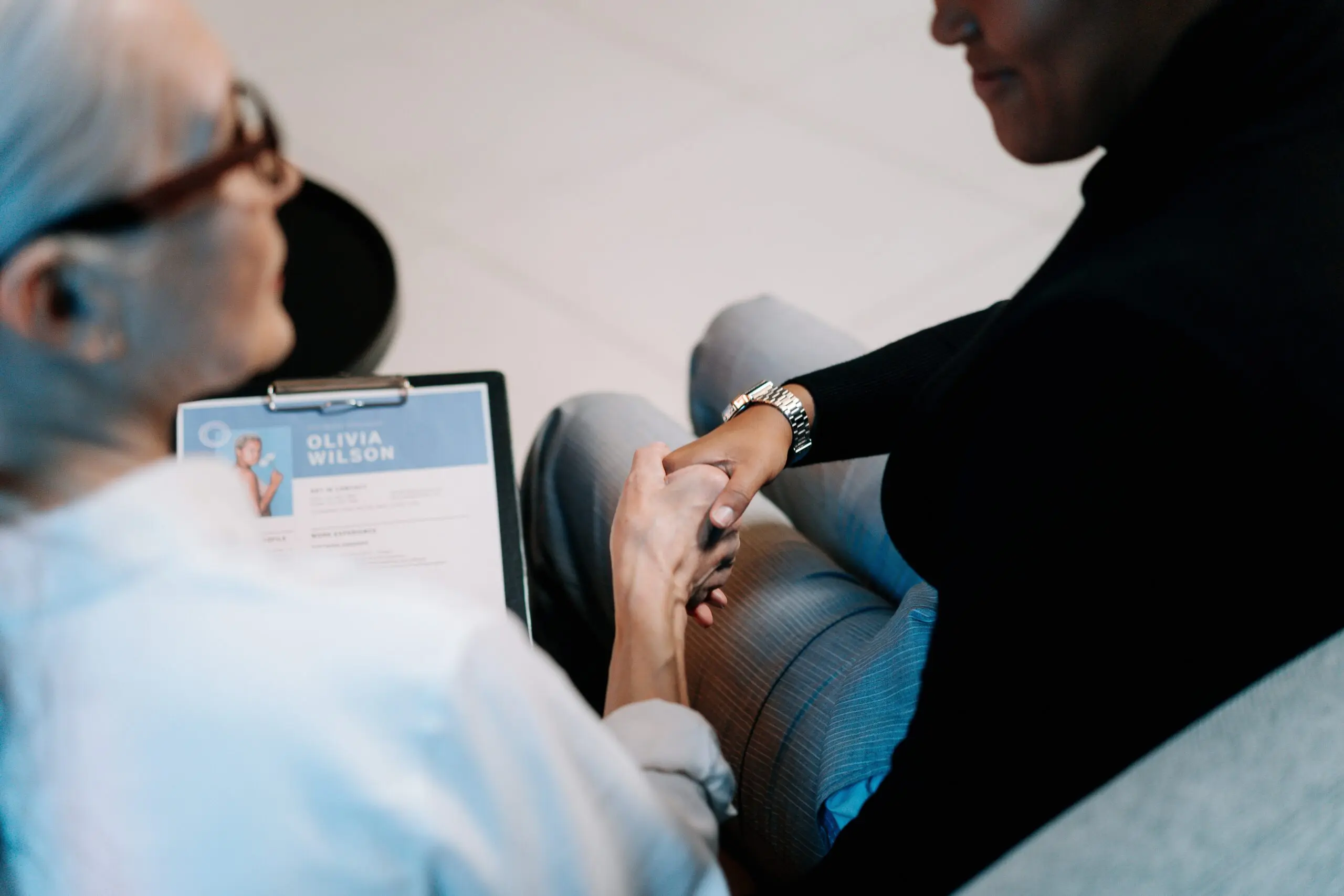 Two people holding hands while sat down, and one has a clipboard with 'Olivia Wilson' on it