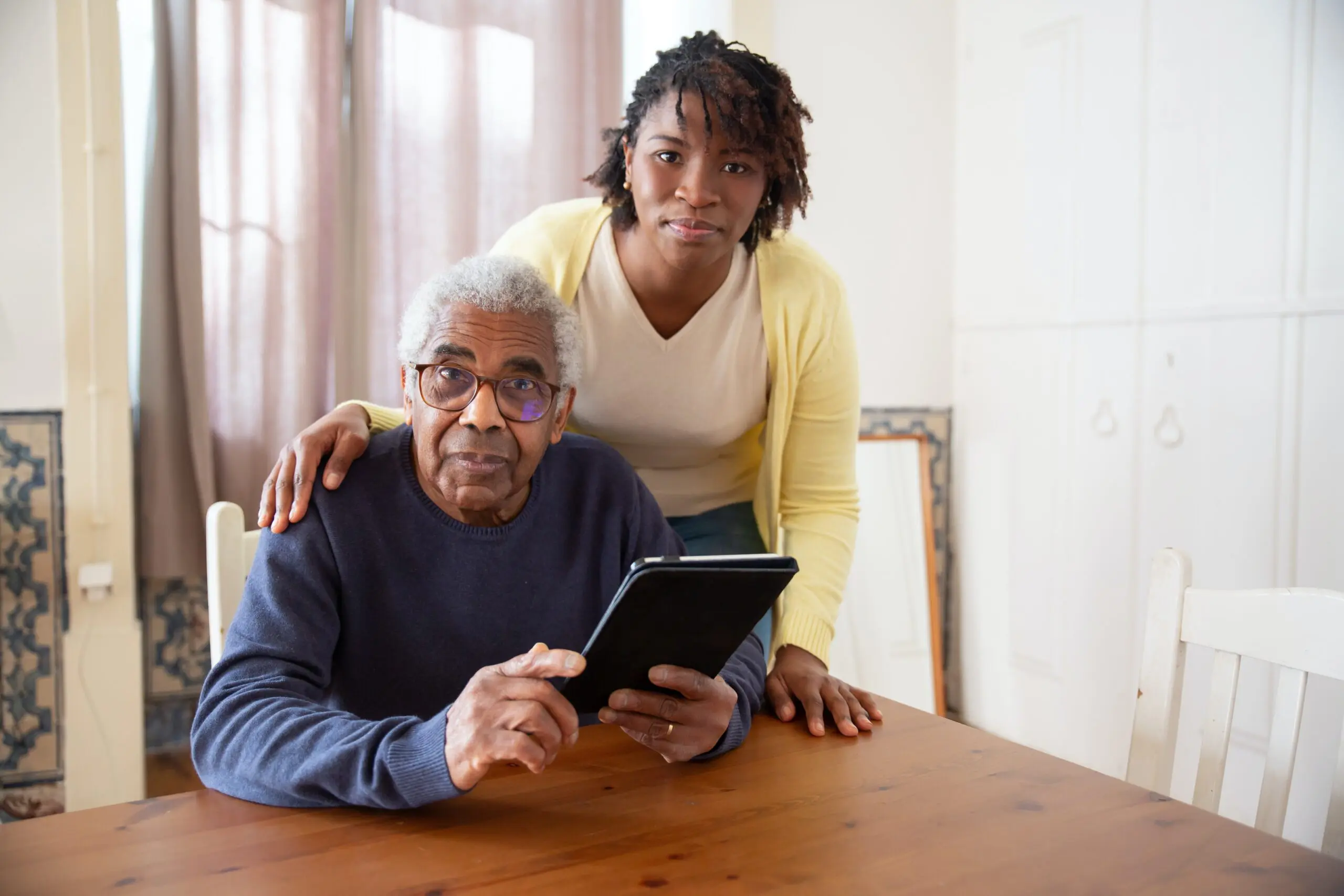 Two people, one sat down, the other with their hand on the shoulder of the first, looking towards the camera