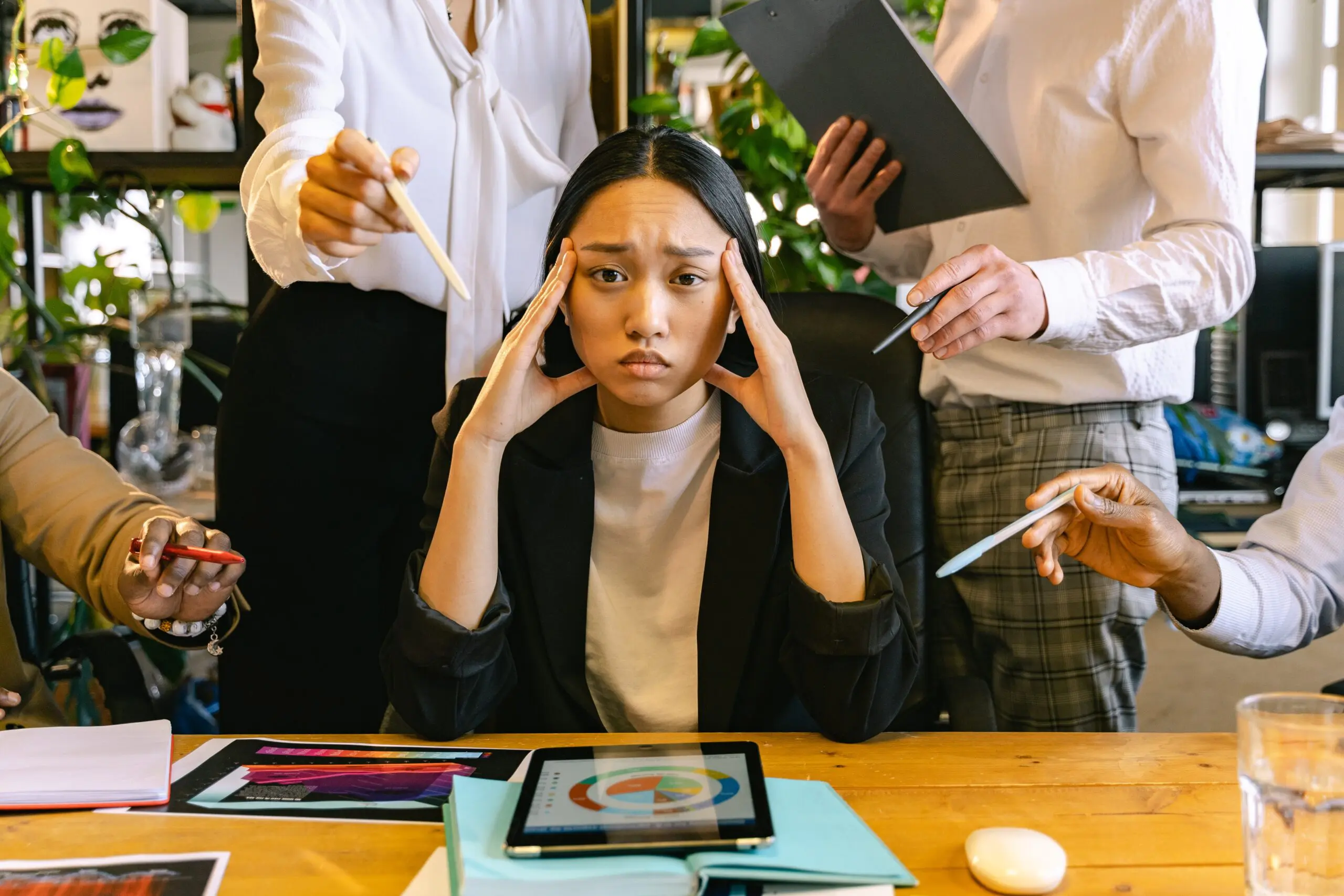 A woman with her head in her hands staring at the camera while several people around her hold out pens