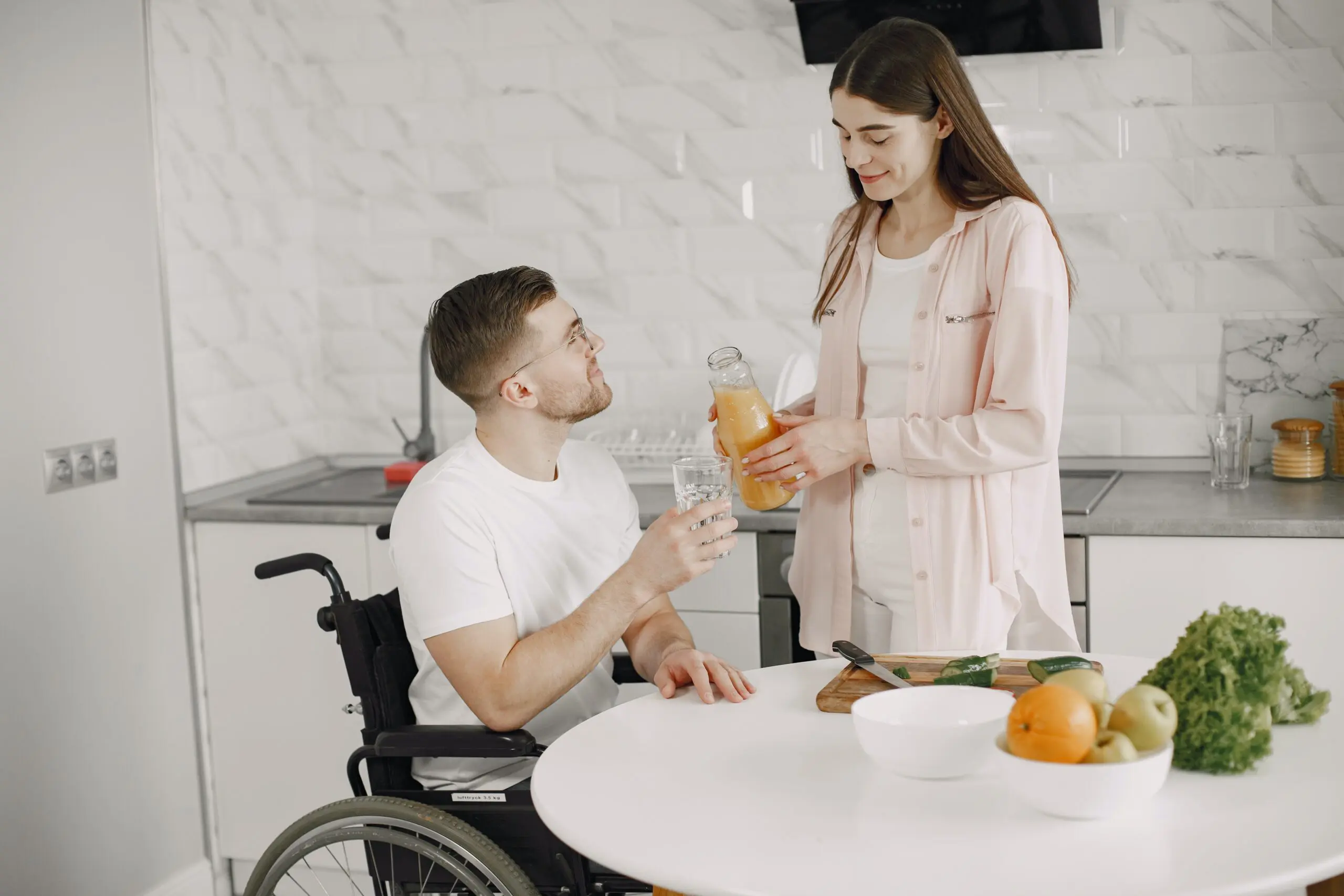 A woman pouring a glass of juice in a kitchen for a man in a wheelchair