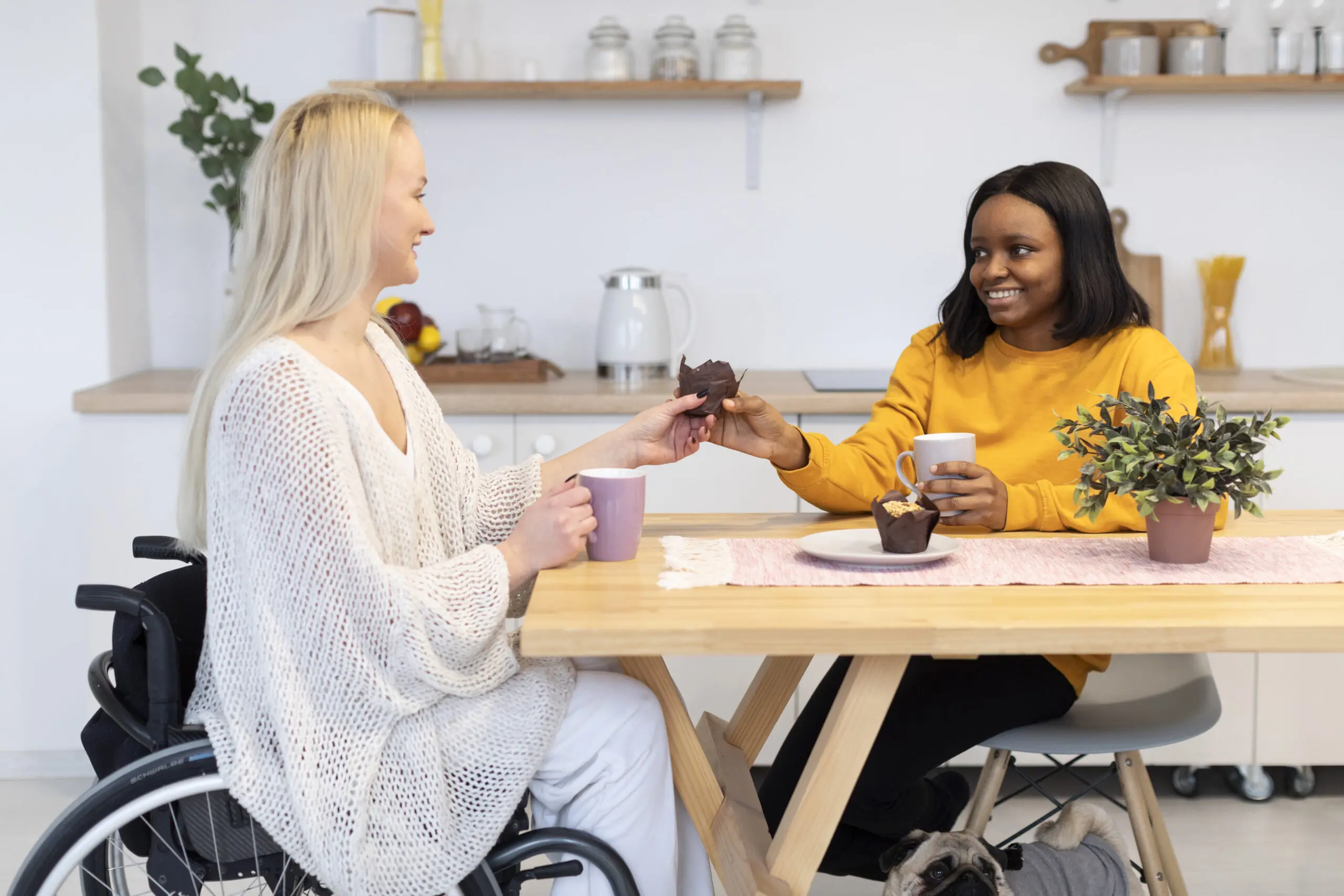 white woman in a wheelchair and black woman sat at kitchen table together, chatting and eating muffins