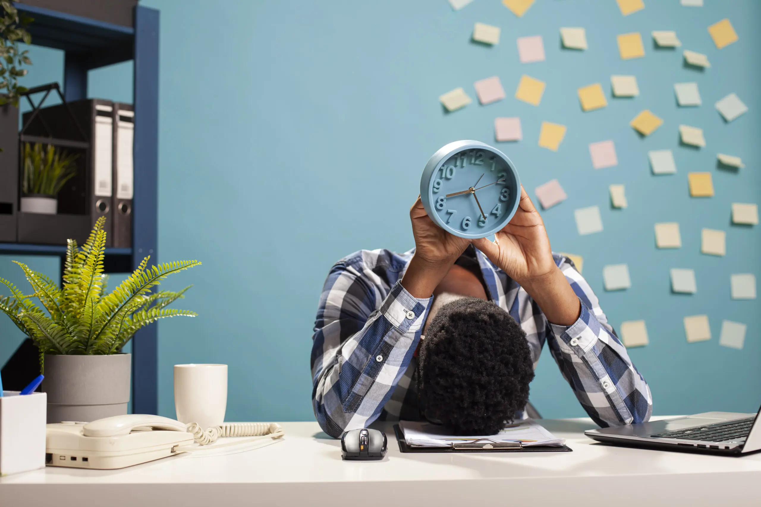 Exhausted male manager resting his head on table and carrying an analogue clock, counting down to end of workday. African american businessman collapsed on office desk, holding alarm clock above him.