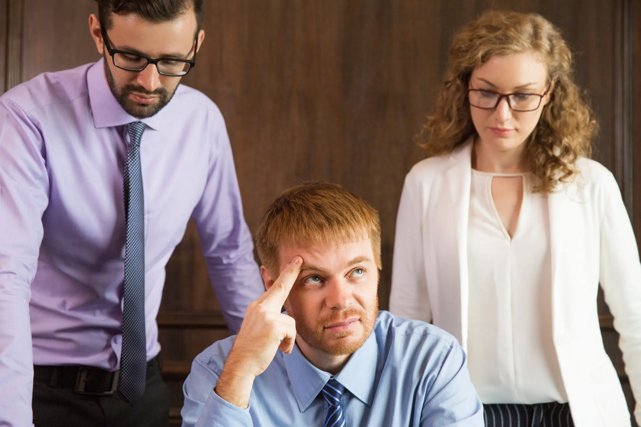 Close up portrait of three care workers thinking. One man is sitting and leaning his head on hand. Other male and female colleagues are standing behind. All are clashing with each others personalities