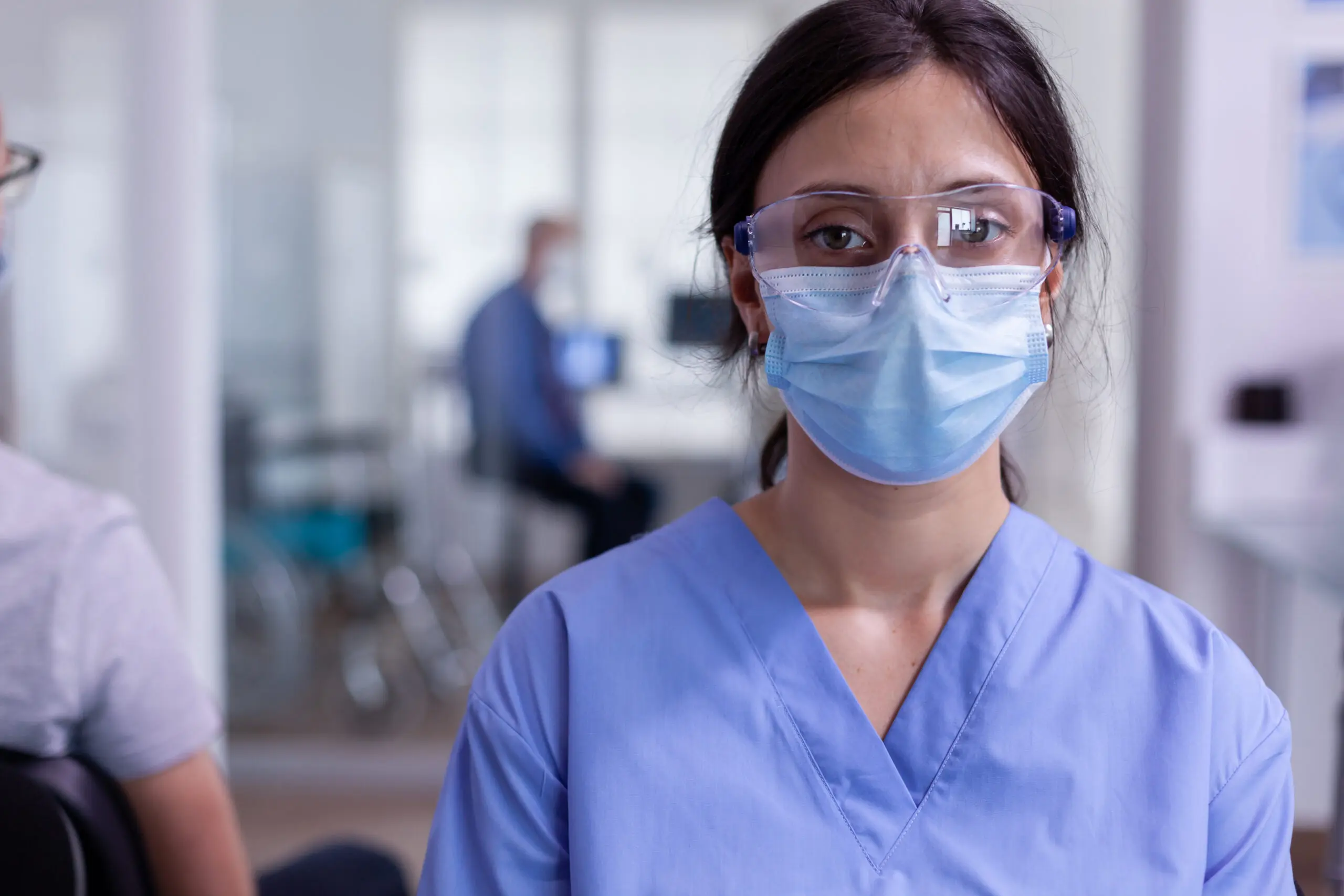 Close up of tired nurse with protection mask against coronavirus outbreak looking at camera sitting in hospital waiting room. Patient with doctor discussing in hospital examination room.