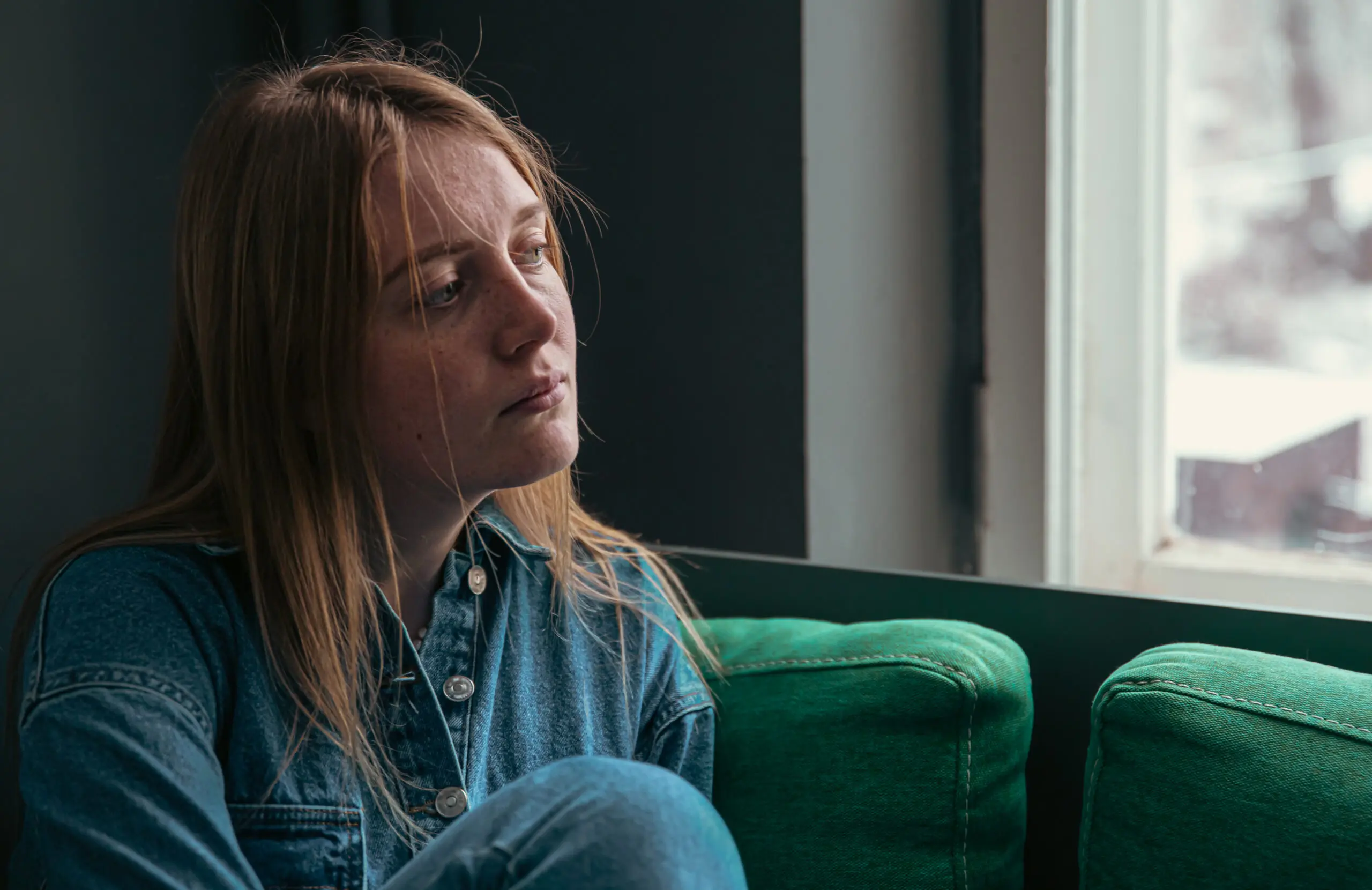 young blonde woman sitting on her sofa alone, looking out the window. She is feeling lonely but doesn't know who to turn to for support