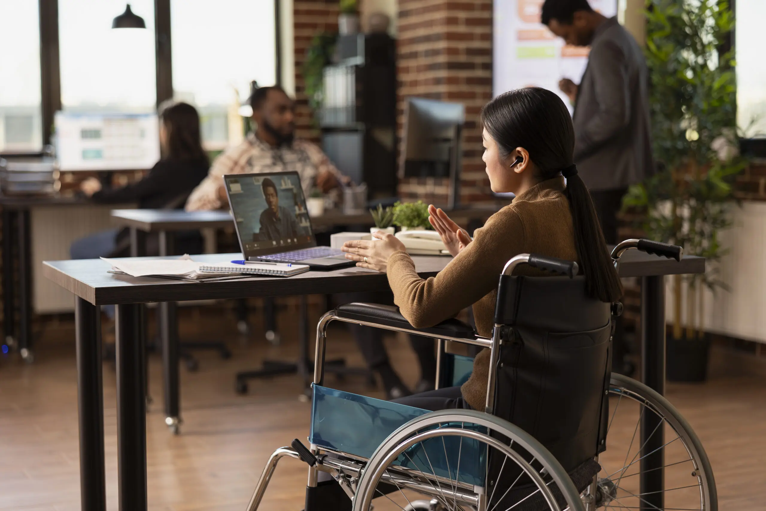 Female manager with disability sits in modern office, leading a virtual meeting with a remote colleague. Asian woman in wheelchair uses her laptop to engage in video call with african american client