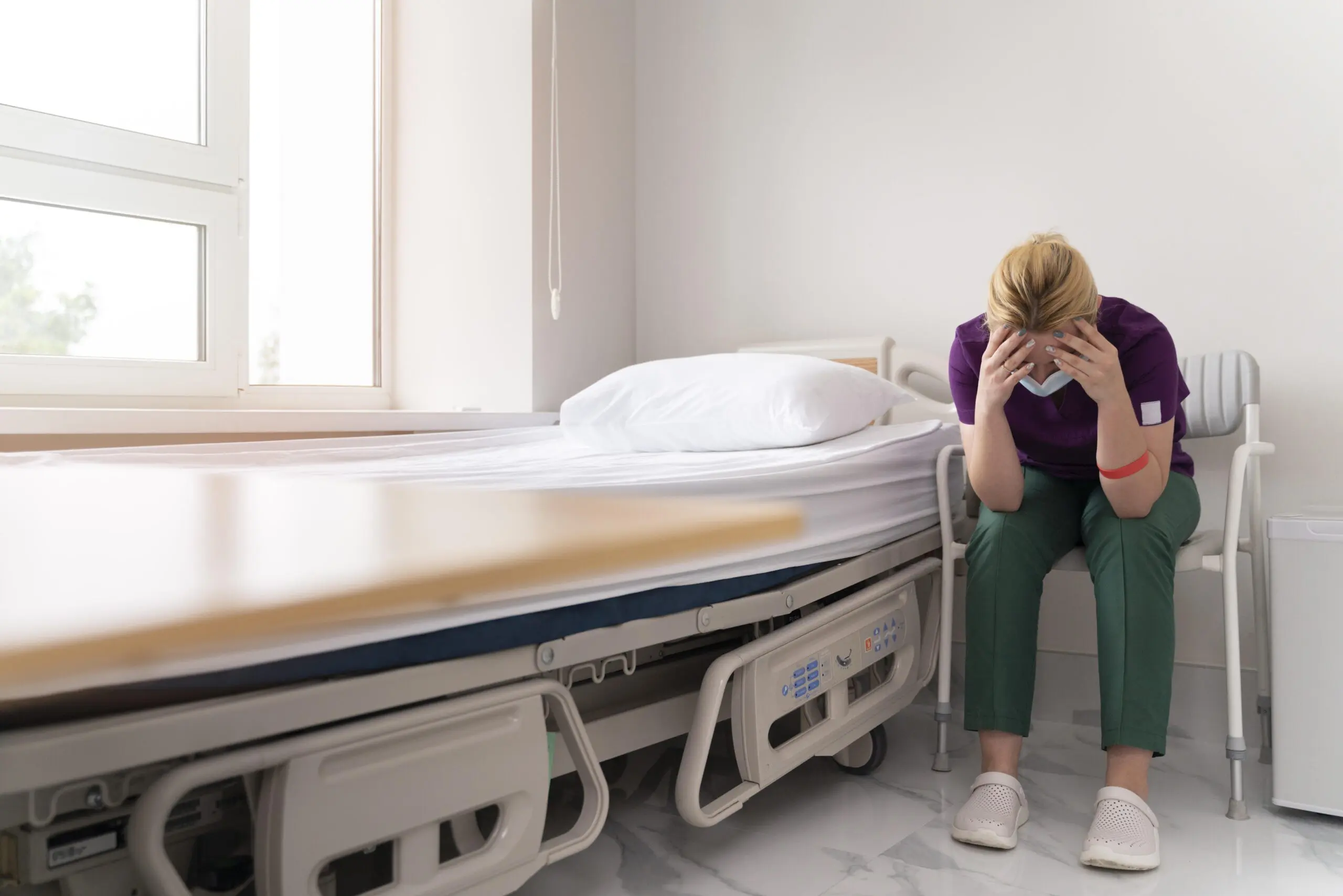 support worker with her head in her hands, in a mental health ward next to an empty bed